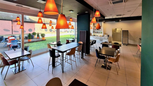 A modern restaurant with multiple tables and chairs, large windows, and hanging orange lamps. - ibis Delemont Delsberg Hotel (Delemont)