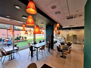 Un restaurante moderno con varias mesas y sillas, ventanas amplias y lámparas naranjas colgantes.