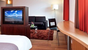 A hotel room with a bed, a TV on a wooden stand, a black sofa, a small table with a vase, a floor lamp, and a wooden desk. - ibis Delemont Delsberg Hotel (Delemont)