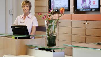 Un mostrador de recepción con una computadora, un florero con flores y dos pantallas mostrando información.