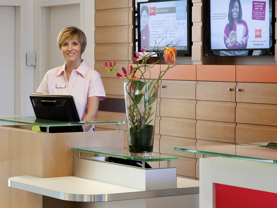 A reception desk with a computer, a vase with flowers, and two screens displaying information.