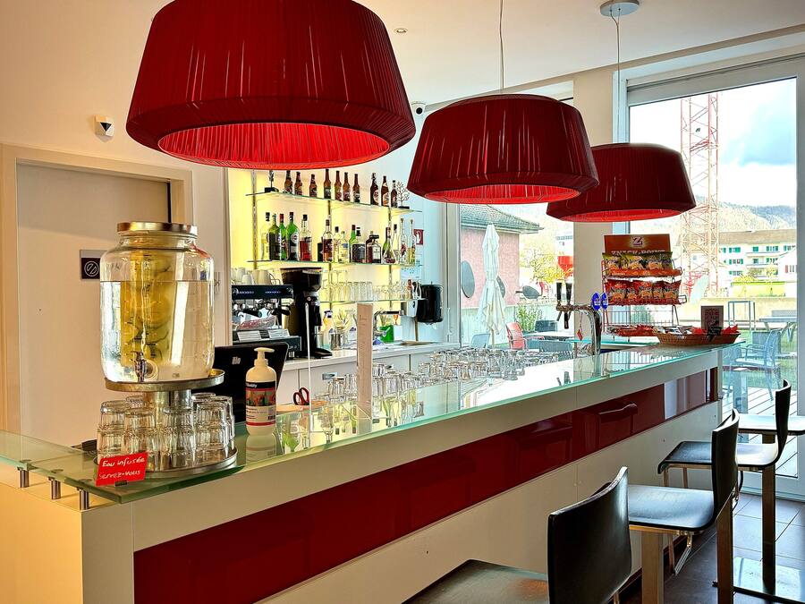 Zona de bar con lámparas colgantes rojas, un expositor de vidrio con botellas y vistas al exterior.