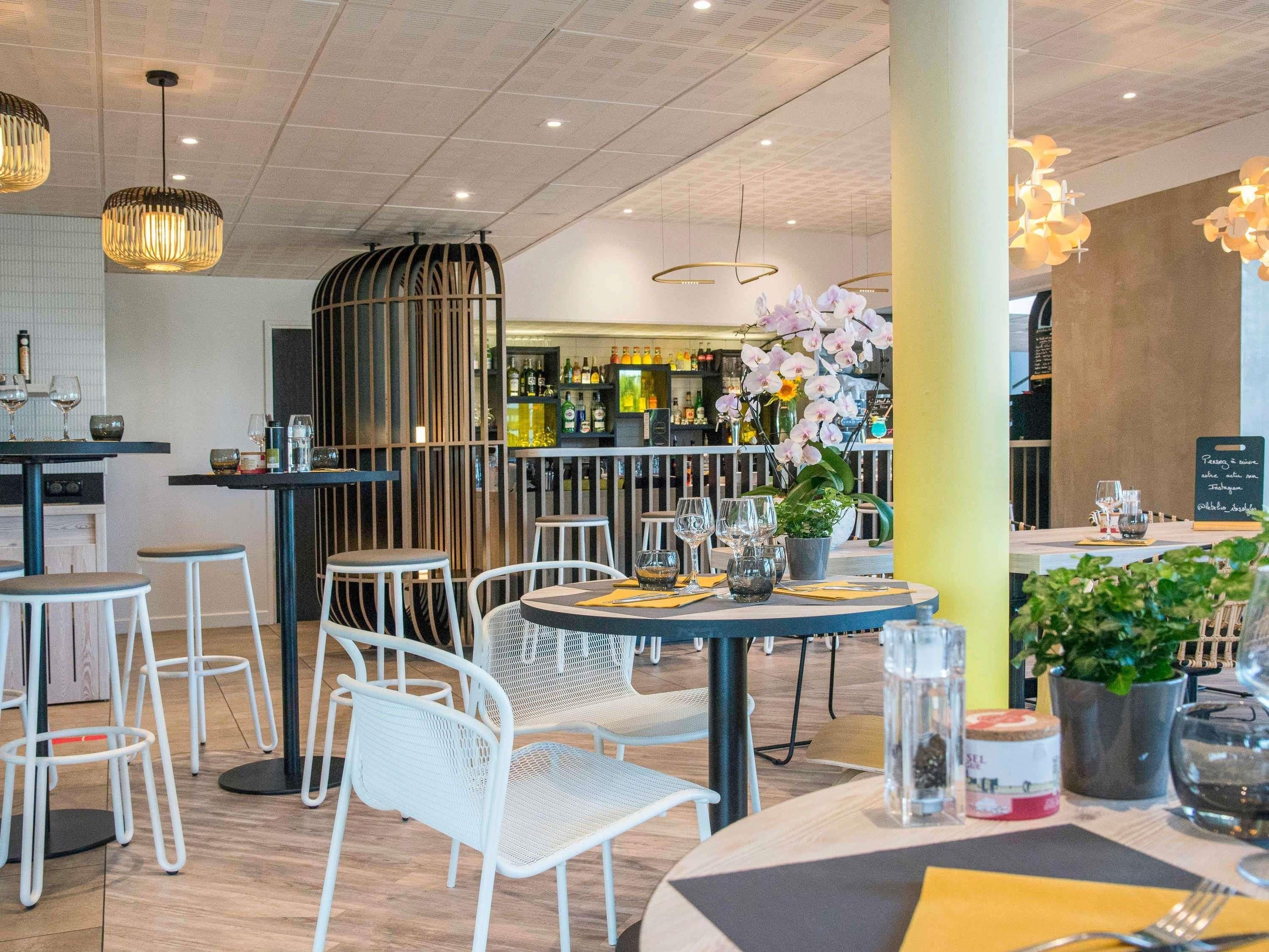 Un interior de restaurante moderno con mesas redondas, sillas blancas, lámparas colgantes y una zona de bar al fondo.