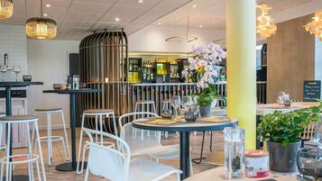 Un interior de restaurante moderno con mesas redondas, sillas blancas, lámparas colgantes y una zona de bar al fondo.