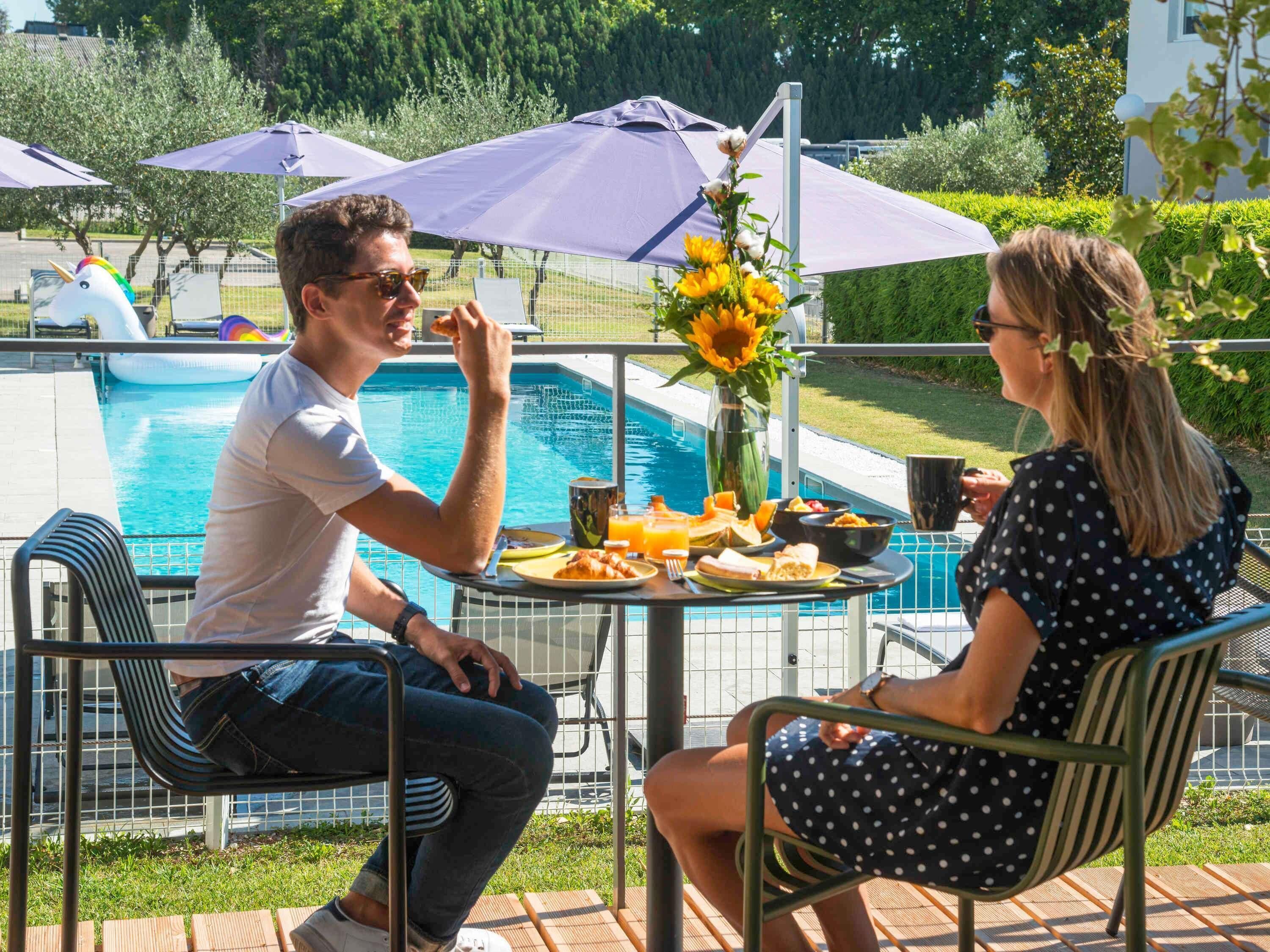 A poolside dining area with a table set for two, surrounded by umbrellas, and a pool in the background.