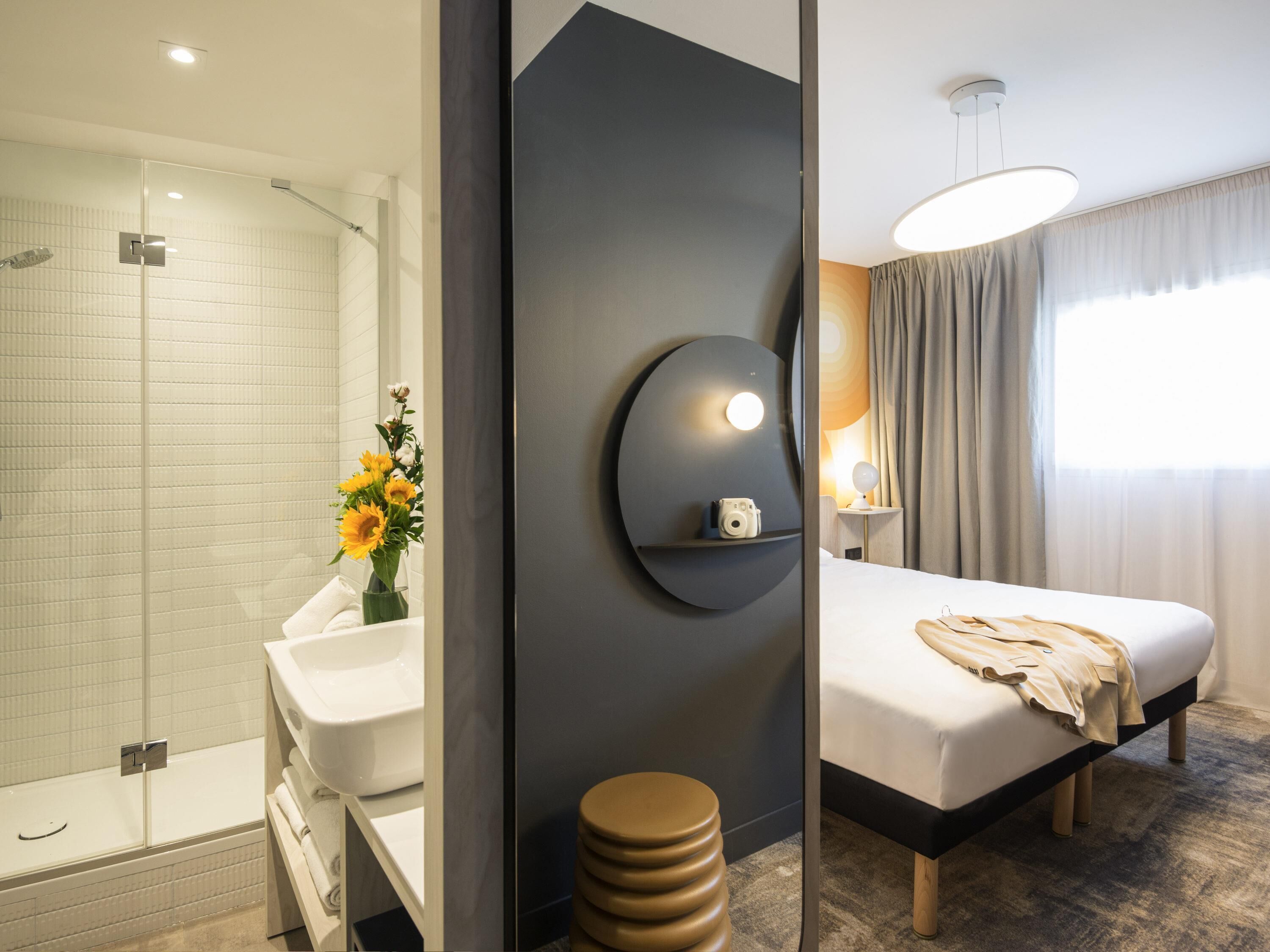 Habitación de hotel con ducha, lavamanos, espejo, cama, mesita de noche y estante en la pared.
