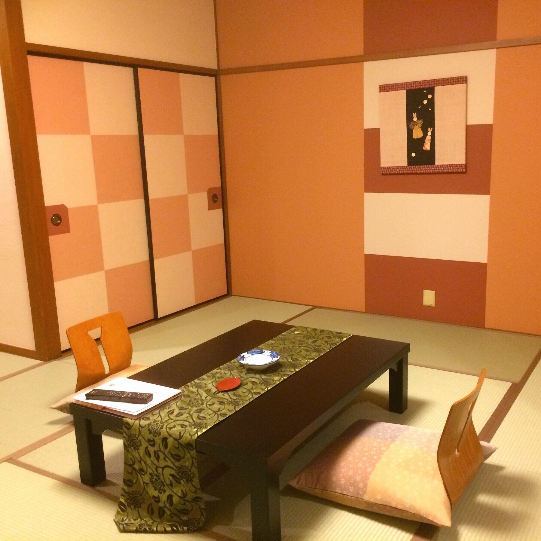A traditional Japanese room with tatami flooring, a low table with a green patterned tablecloth, a cushion, and a framed picture on the wall.
