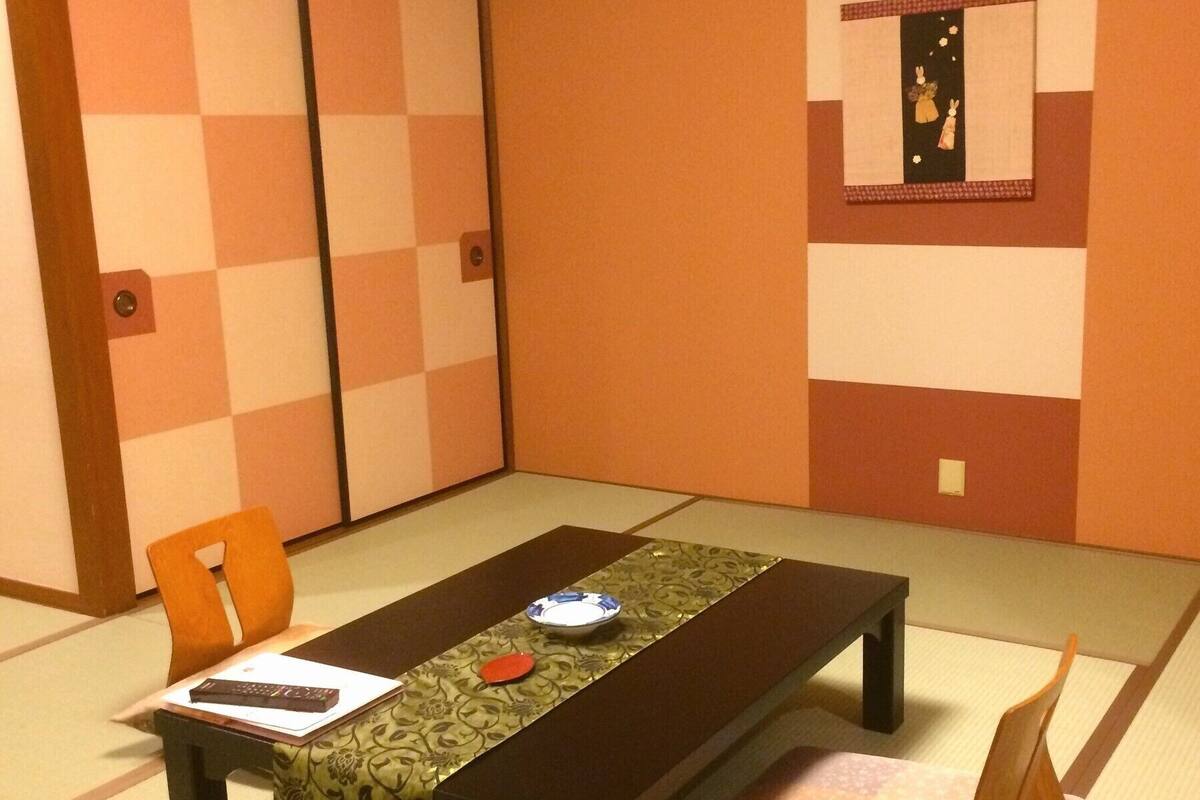 Ein traditioneller japanischer Raum mit Tatami-Boden, einem niedrigen Tisch mit grün gemustertem Tischläufer, einem Kissen und einem gerahmten Bild an der Wand.