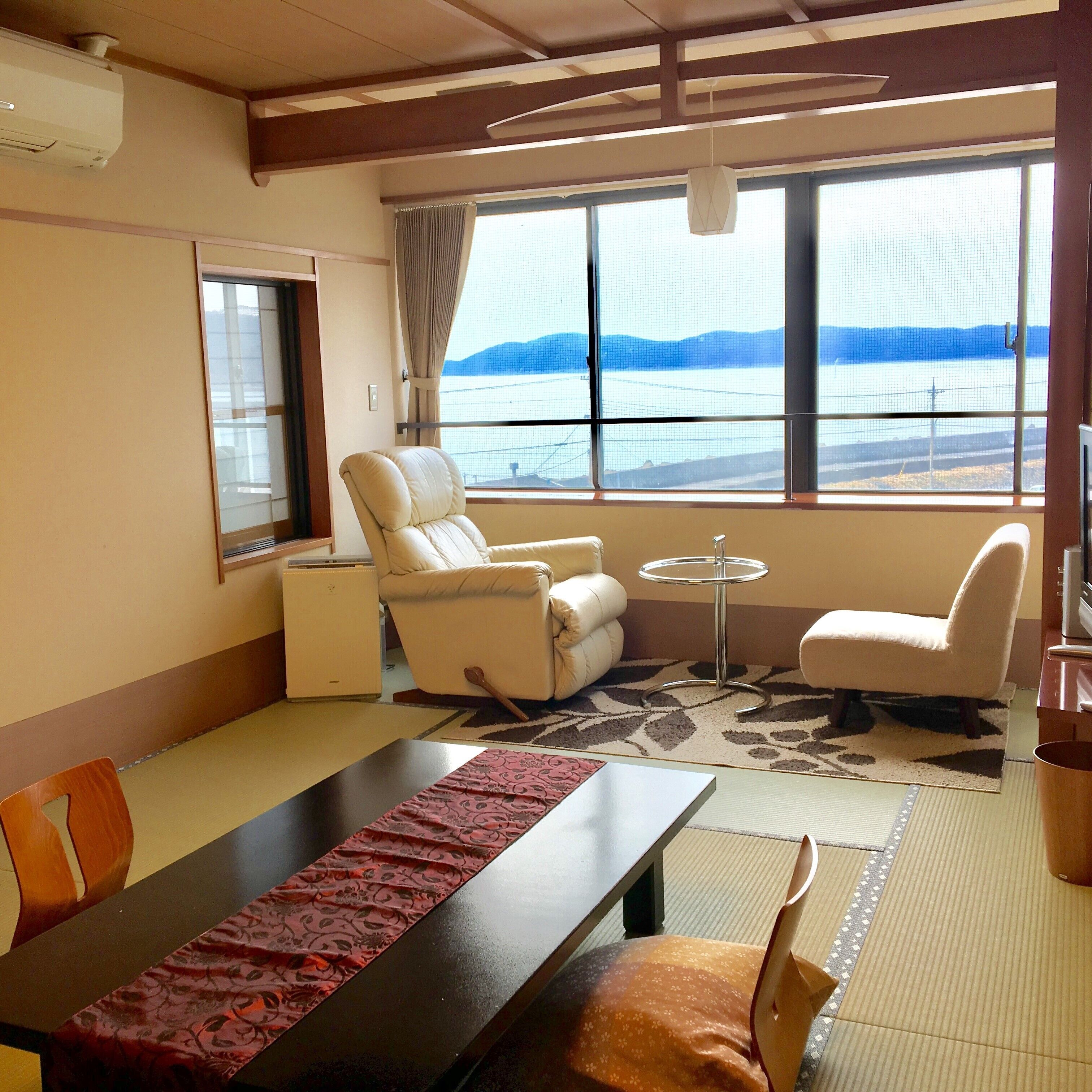 A room with a dining table, two chairs, a sofa, a small round table, and a view of the sea.