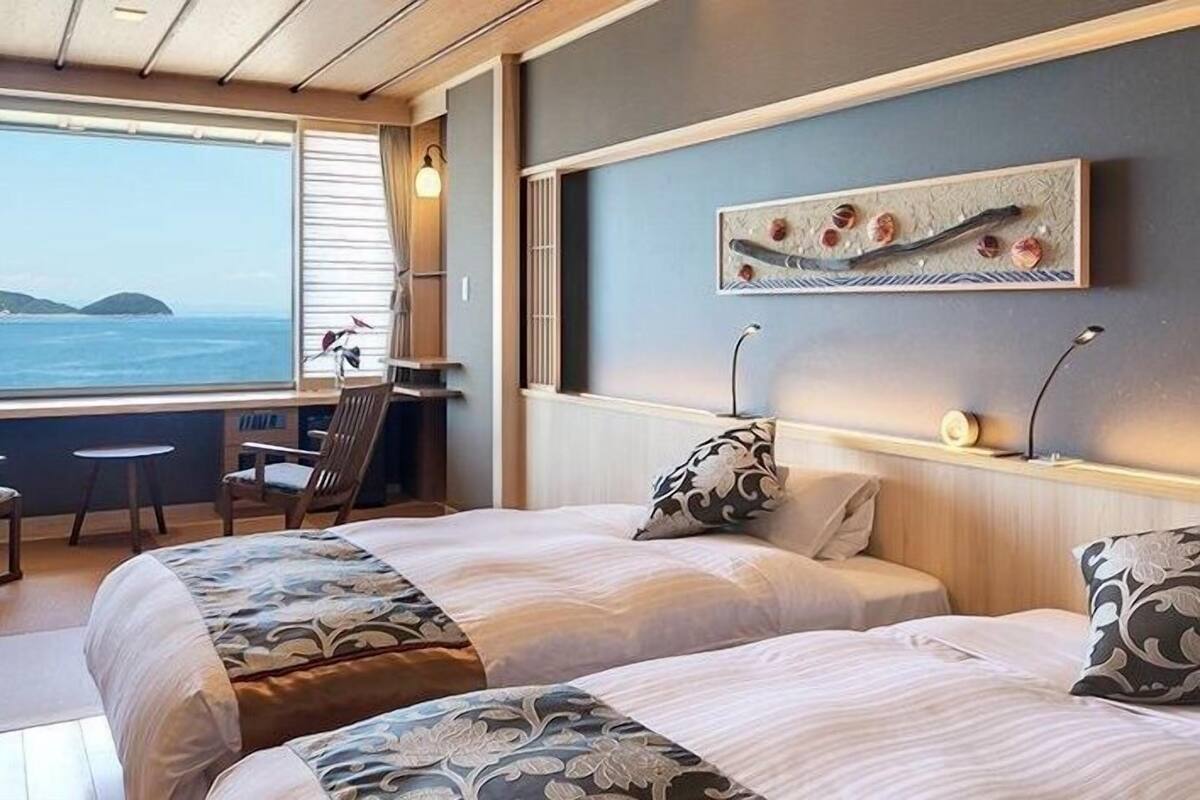 Ein Hotelzimmer mit zwei Betten, einem Schreibtisch, einem Stuhl und einem großen Fenster mit Meerblick.