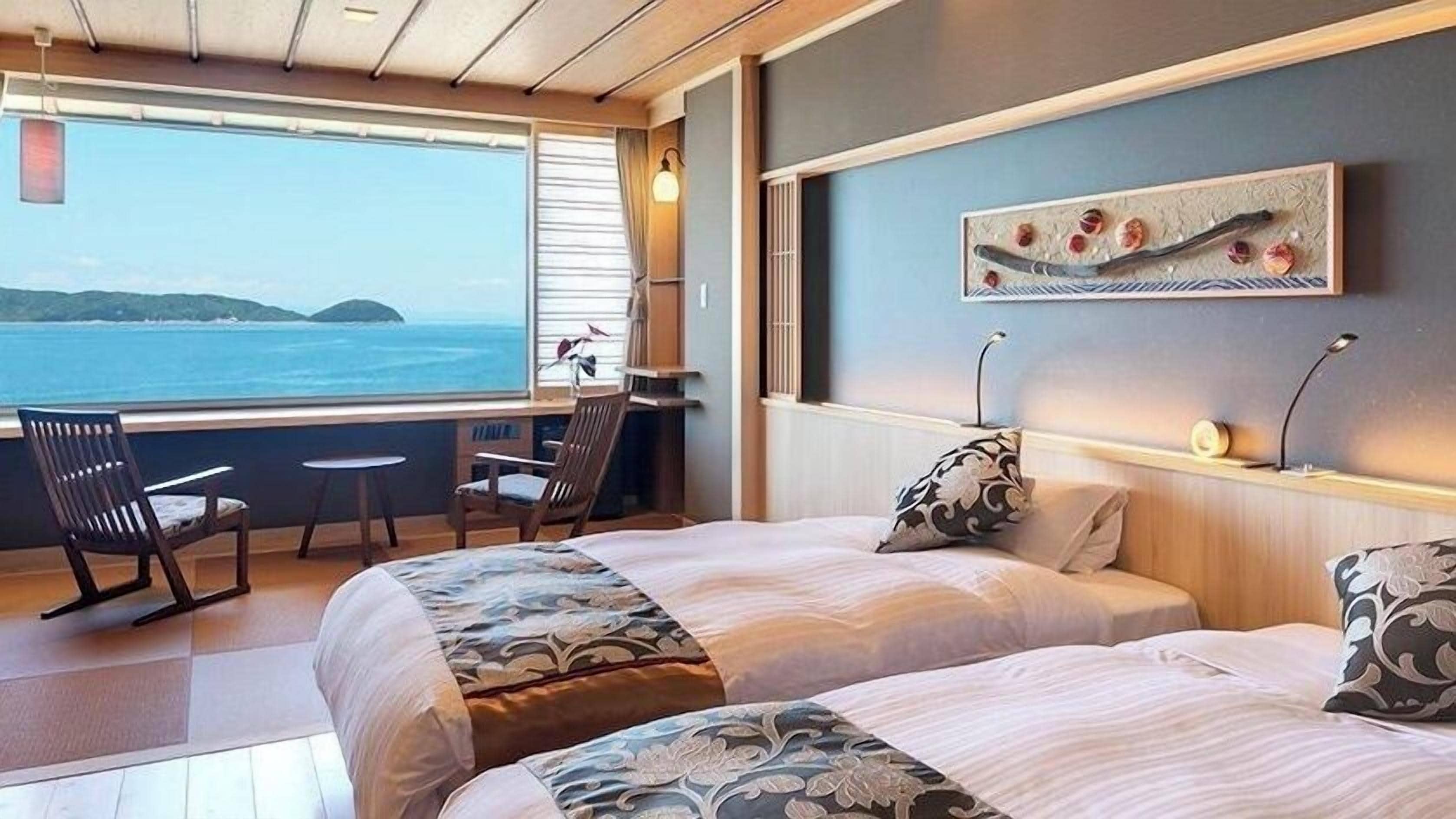 A room with two beds, a desk, and a chair, with a view of the sea and hills.