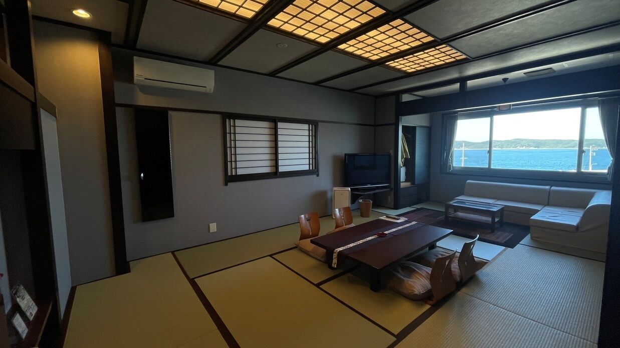 A room with tatami flooring, a television, a sofa, and a view of the sea.