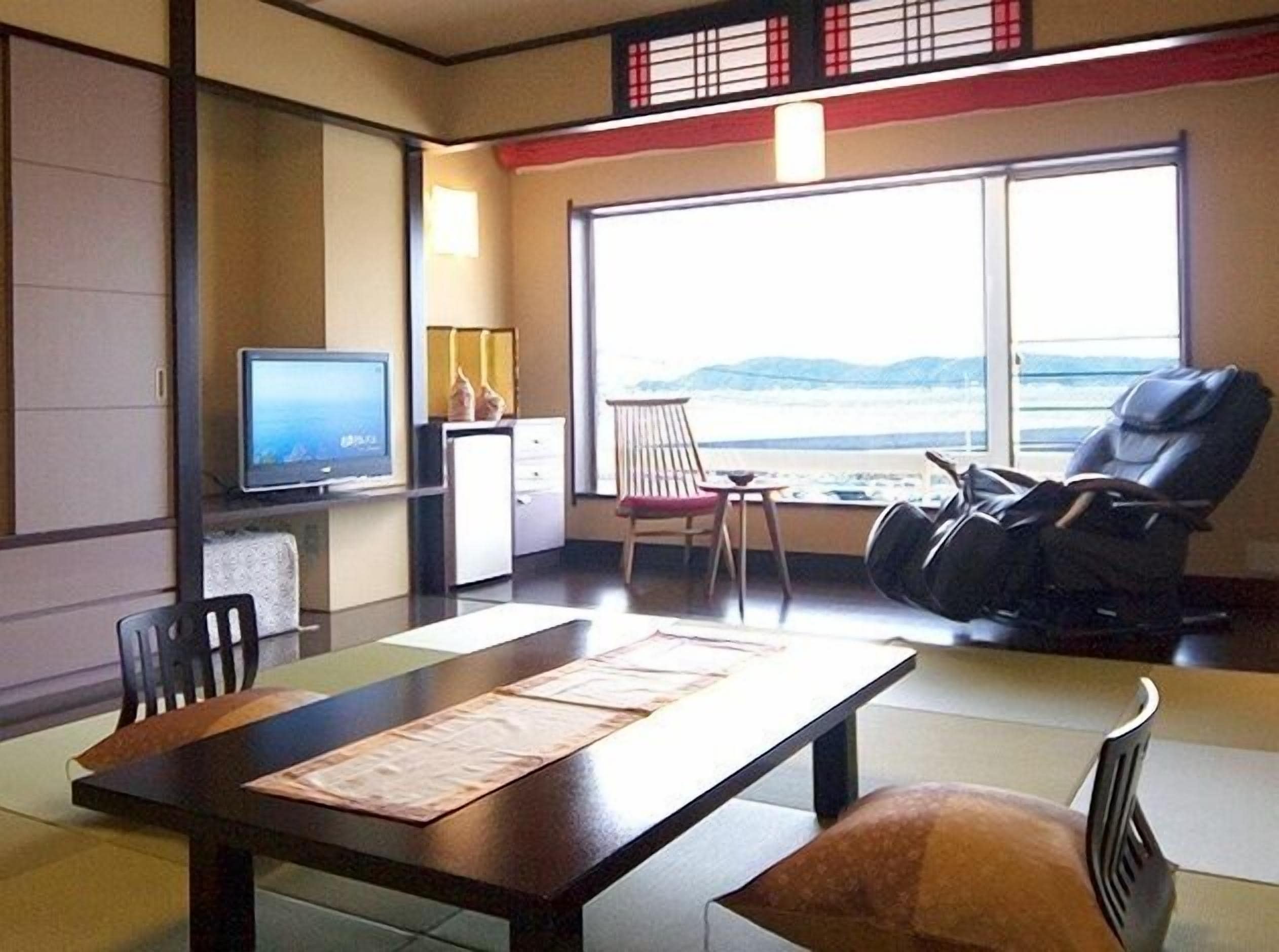 A room with a TV, a massage chair, a dining table with placemats, a window with a view of the sea, and traditional Japanese decor.