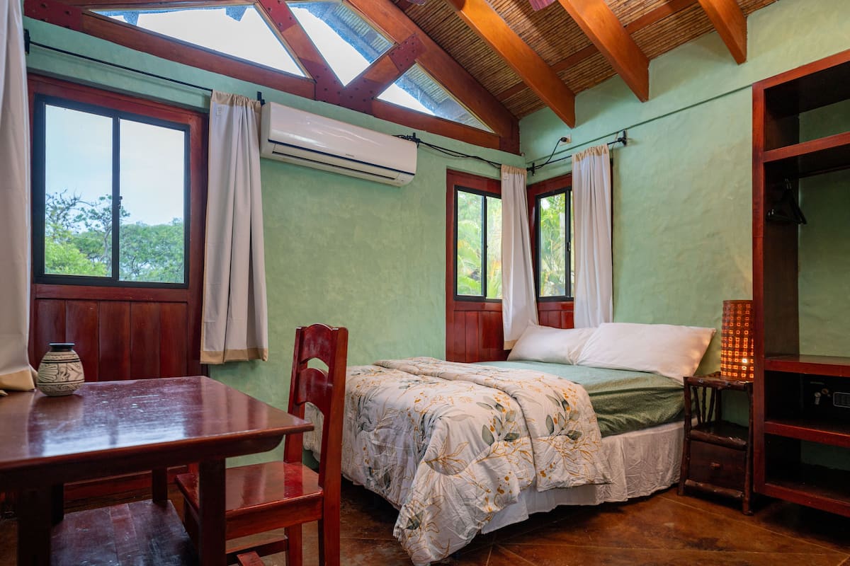 A room with a bed, a wooden table, a chair, and a small table with a vase. There are windows with curtains, an air conditioning unit, and a shelf with a lamp.