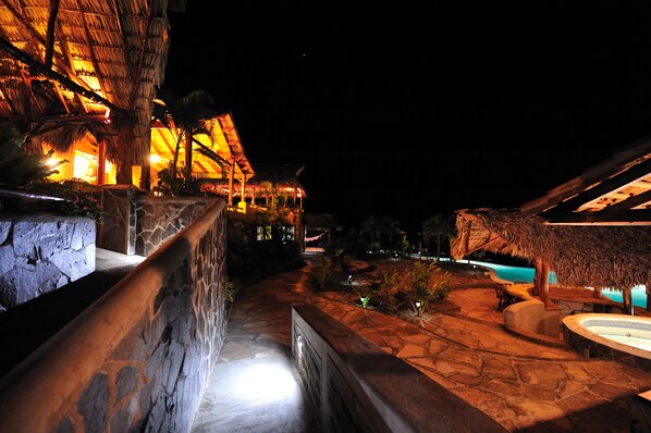 A nighttime view of a resort with stone pathways, thatched-roof structures, and a swimming pool area.