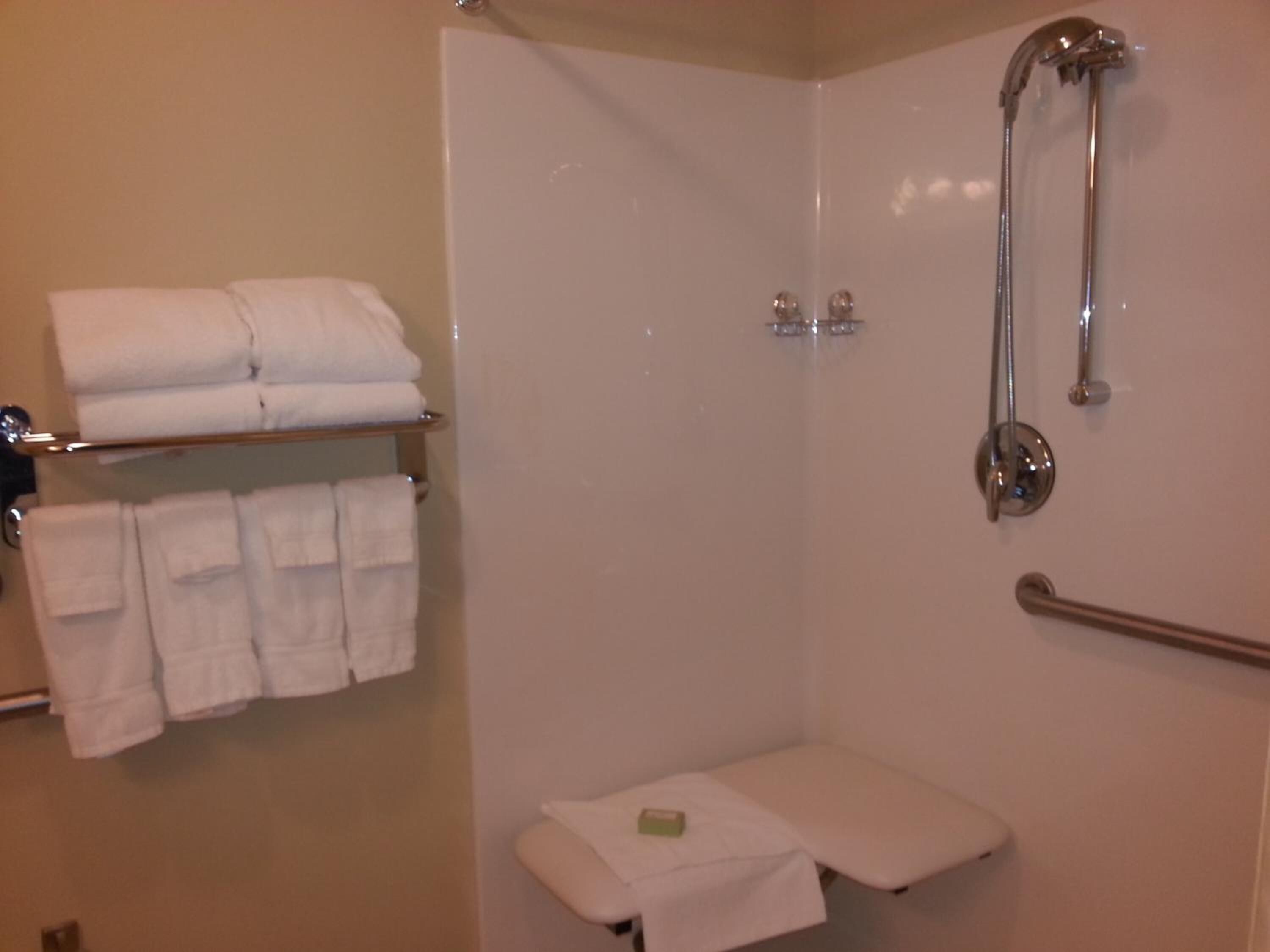 A shower area with a glass door, a towel rack with folded towels, a handheld showerhead, and a small bench with a soap dish.
