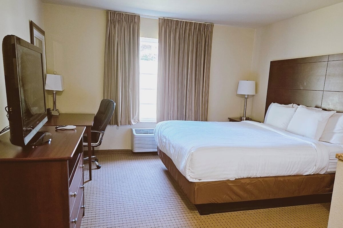 A hotel room with a bed, a desk with a computer, a chair, a TV, and a window with curtains.