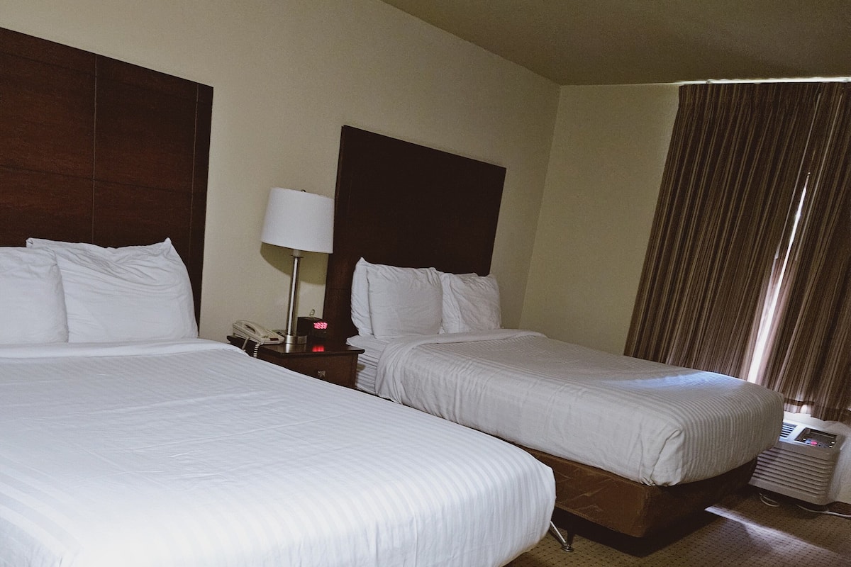A hotel room with two beds, a nightstand with a lamp, and a window with curtains.