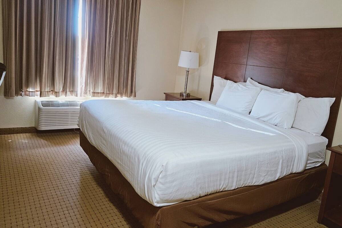 A hotel room with a large bed, a bedside table with a lamp, a window with curtains, and a wall-mounted air conditioner.