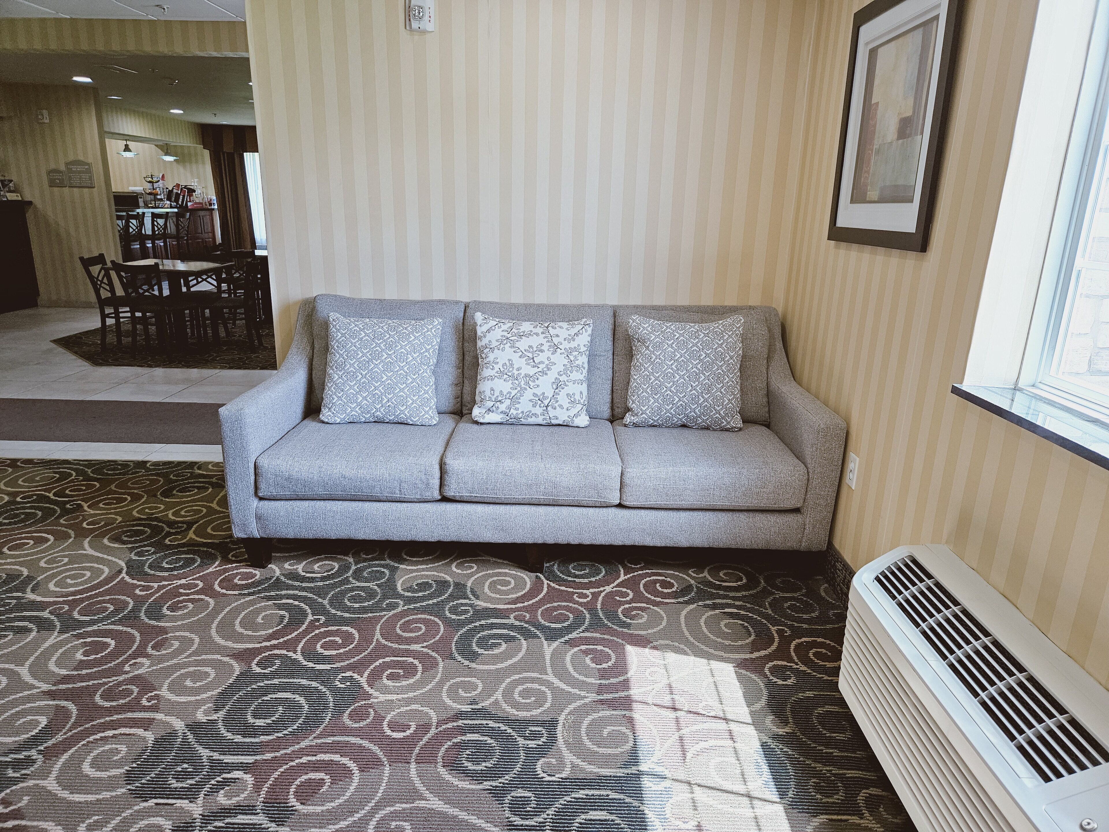 A grey sofa with patterned cushions, a patterned carpet, a window with blinds, and a wall-mounted air conditioner.