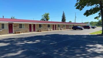 A motel with a red roof and stone facade, featuring multiple rooms with red doors, a parking lot, and a few parked cars.