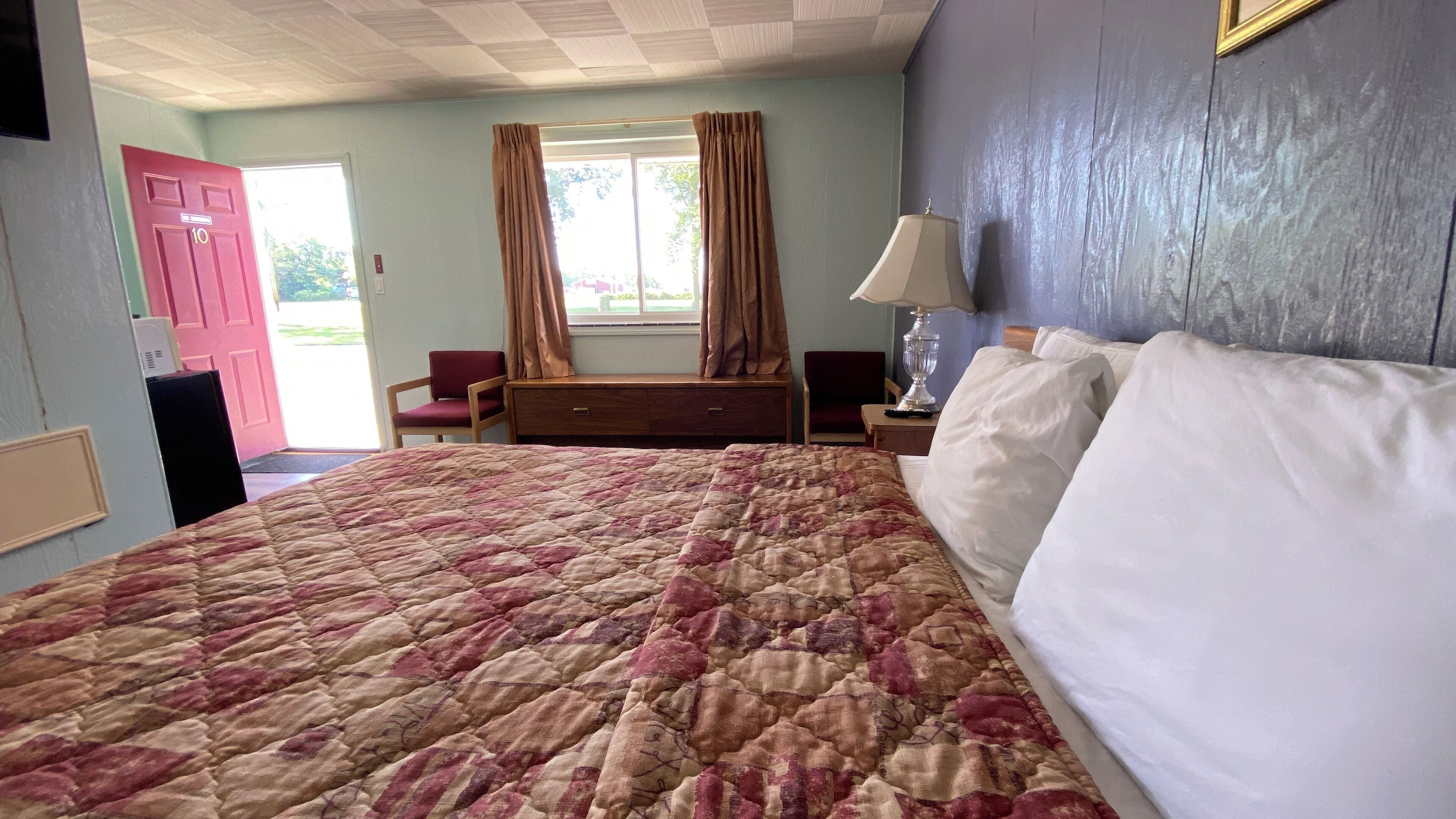 A hotel room with a large bed, two chairs, a nightstand, a lamp, and a window with curtains.