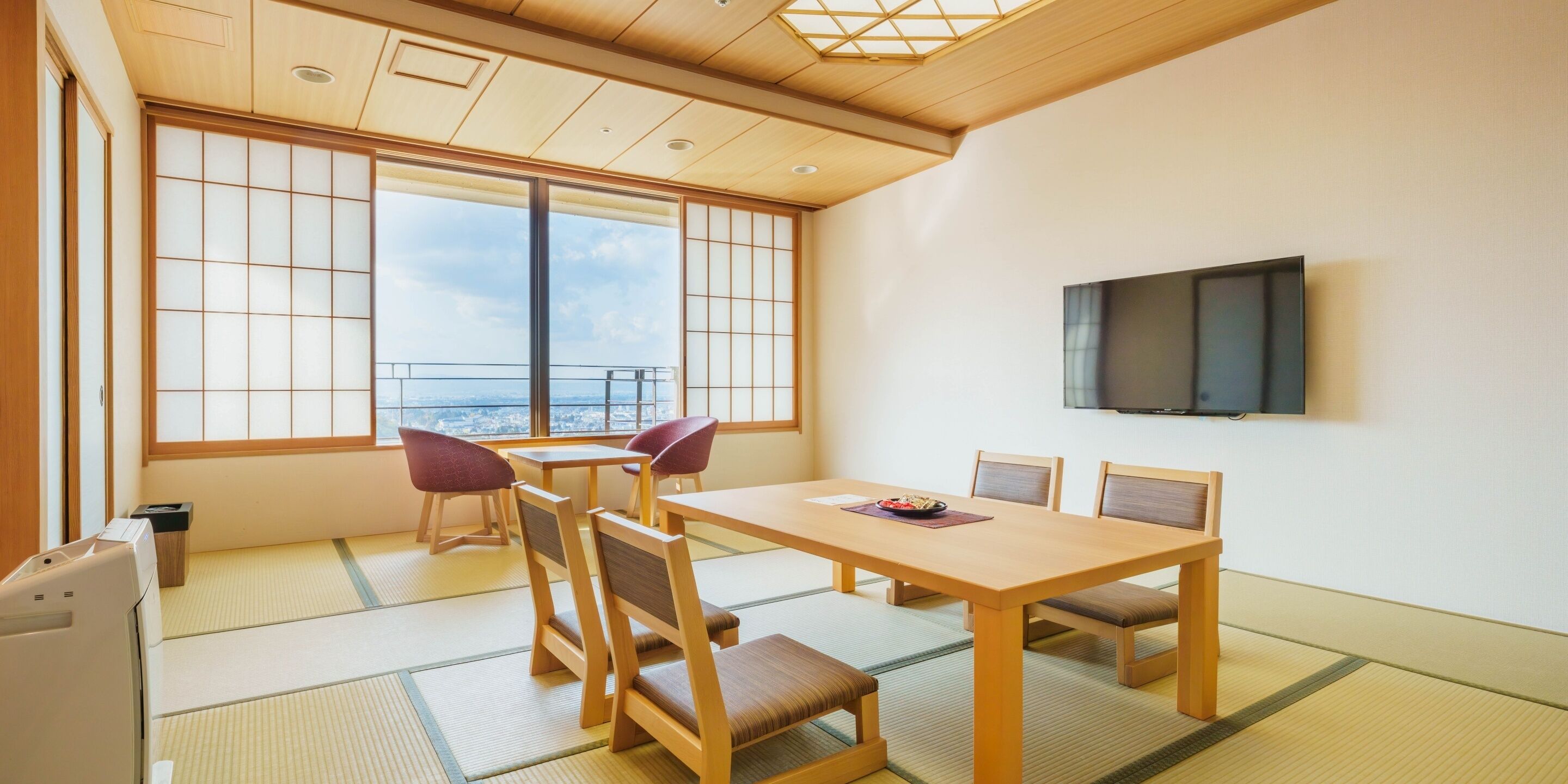 Ein traditioneller japanischer Raum mit Tatami-Boden, einem Holztisch, Stühlen, einem Fernseher und Blick durch große Fenster.