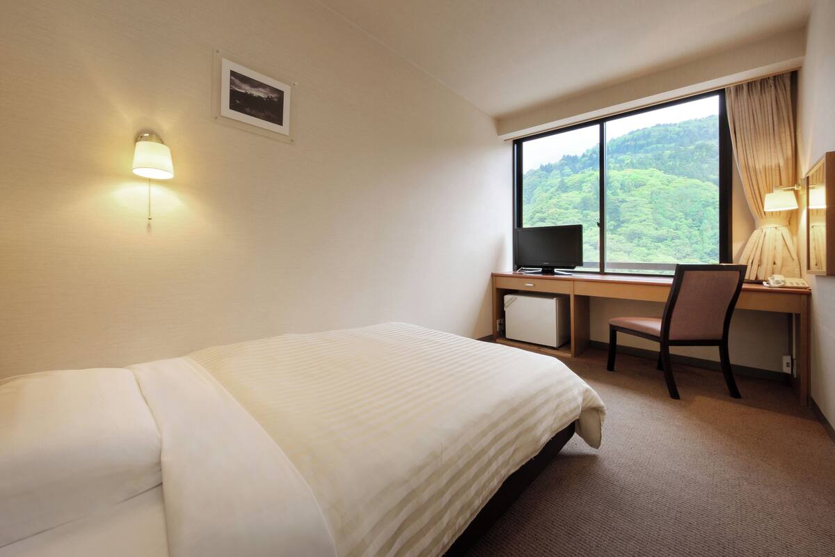Habitación de hotel con cama, escritorio con computadora, silla, refrigerador pequeño y ventana con vista a un bosque.