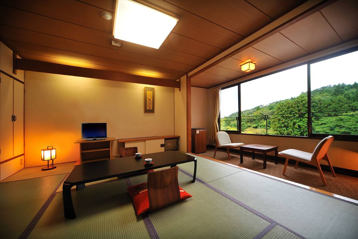 Una habitación japonesa tradicional con suelo de tatami, una mesa baja, sillas, un televisor y vistas a una ladera verde.