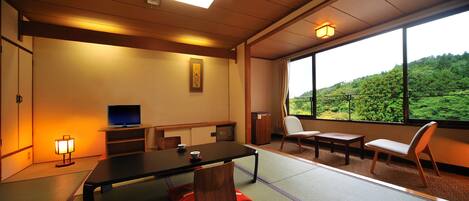 Una habitación japonesa tradicional con suelo de tatami, una mesa baja, sillas, un televisor y vistas a una ladera verde.