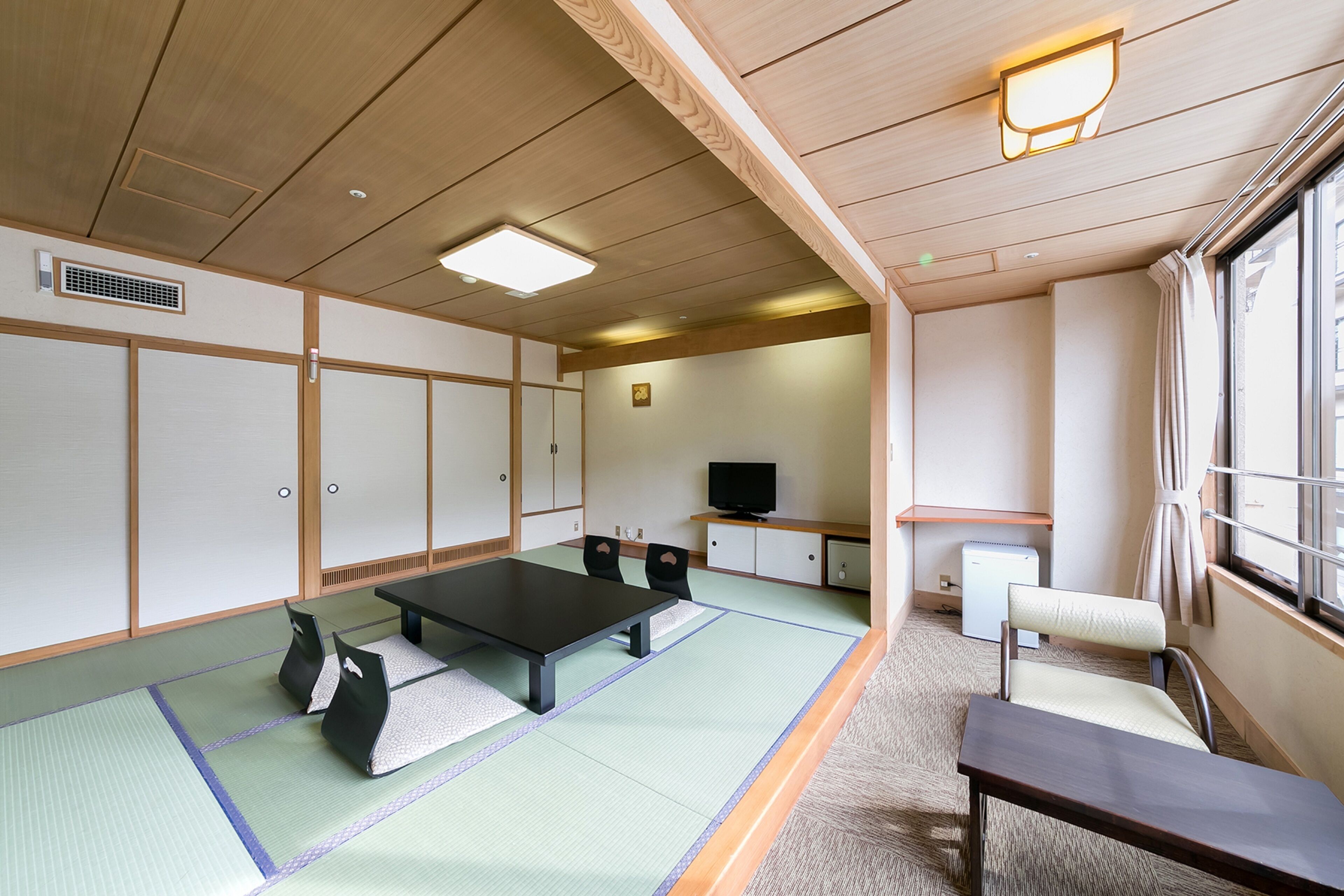 A traditional Japanese room with tatami flooring, a low table, chairs, a television, and a window with curtains.