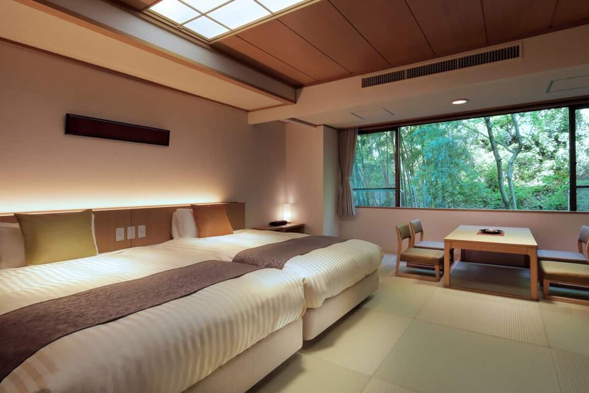 A hotel room with two beds, a table, chairs, a window with a view of trees, and a skylight.