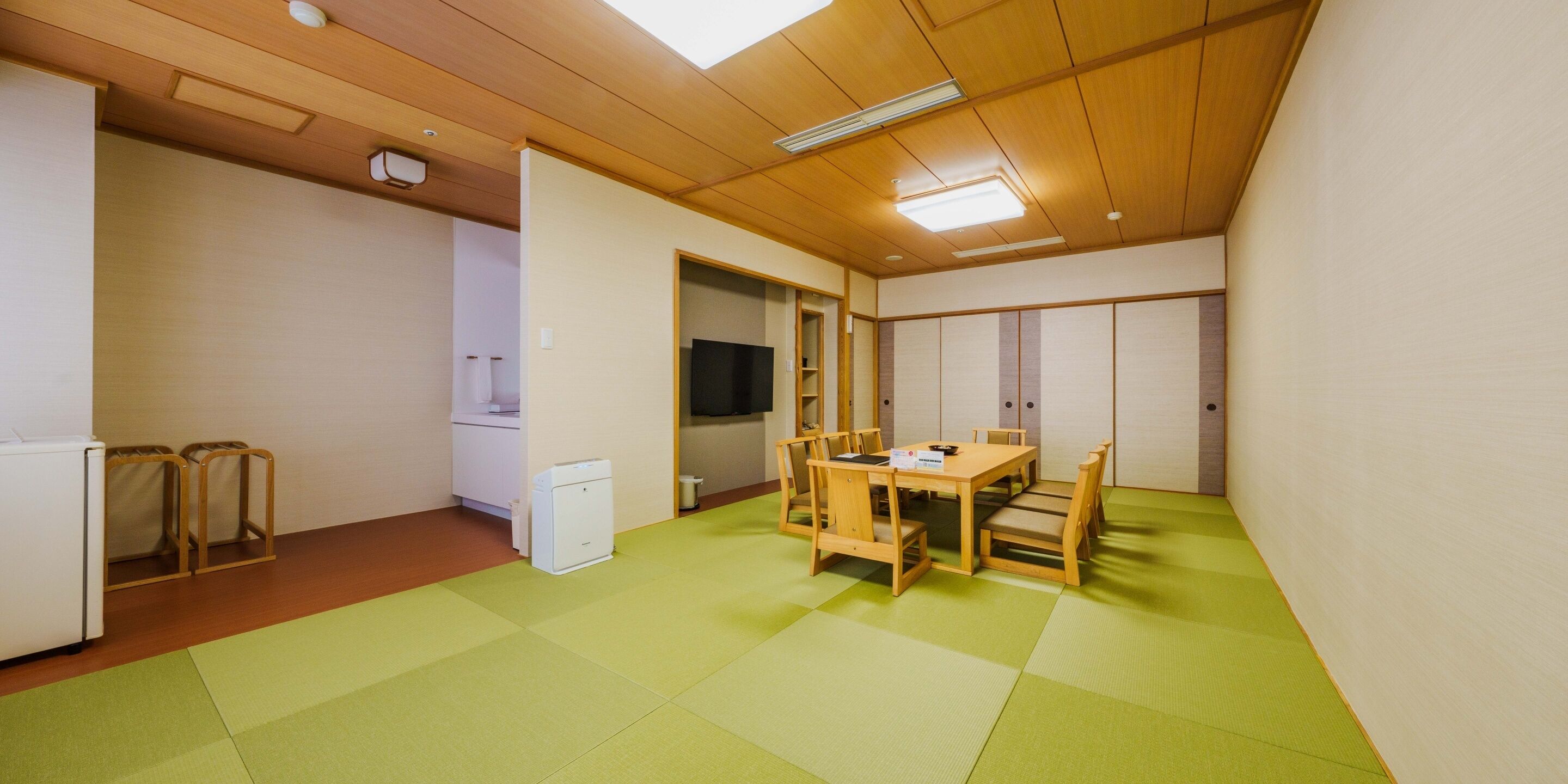 Une pièce avec des tatamis, une table en bois, des chaises, un téléviseur et un réfrigérateur.
