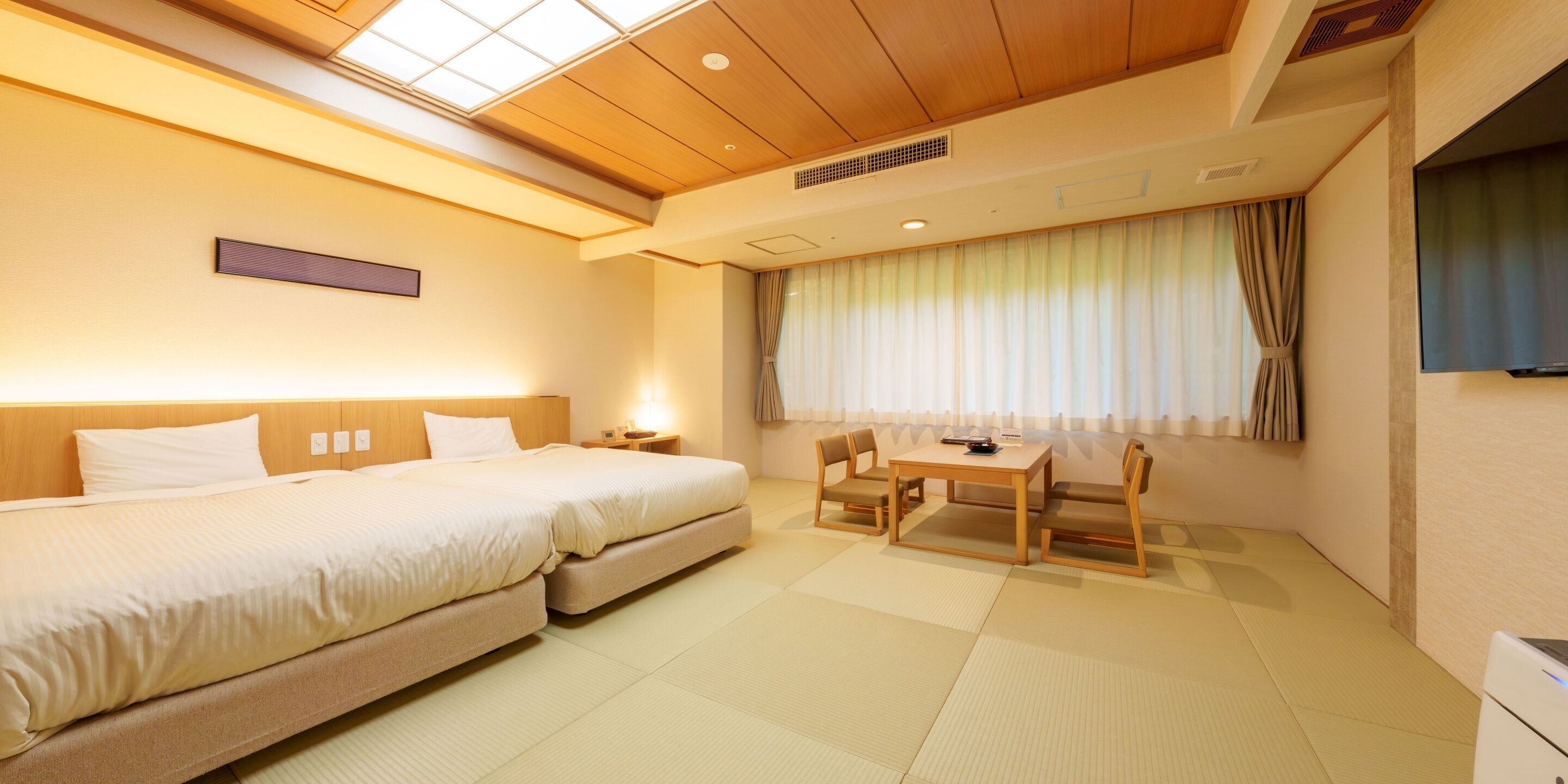 A hotel room with a bed, two bedside tables, a small table with chairs, a TV, and a window with curtains.