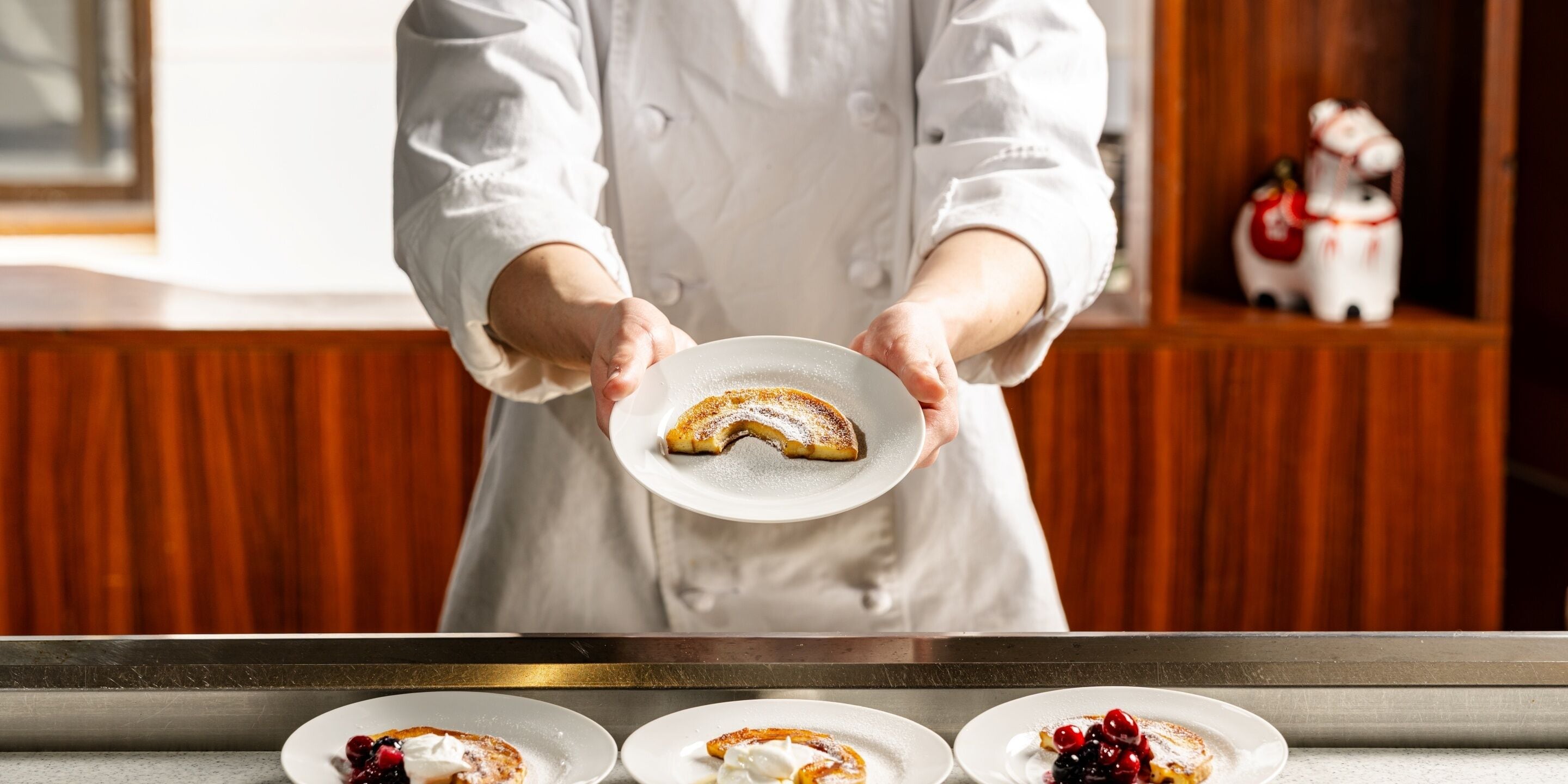 Une personne vêtue d’une blouse de chef blanche, tenant une assiette sur laquelle se trouve un dessert.