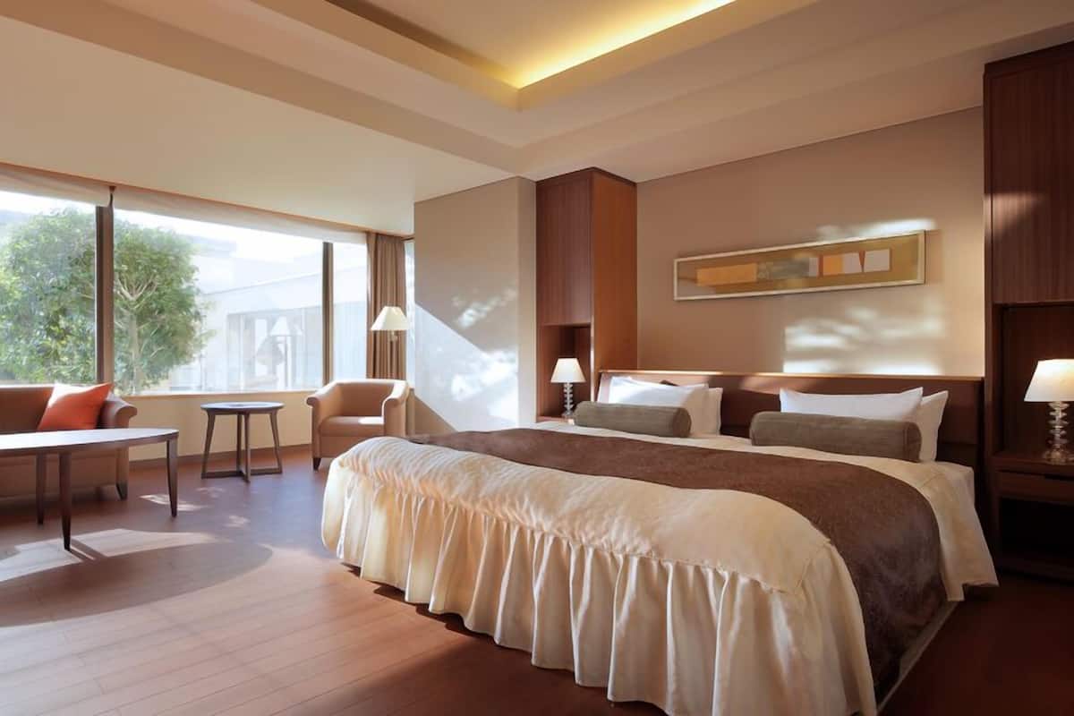 A spacious hotel room with a large bed, a seating area with a table, and a view of greenery outside.