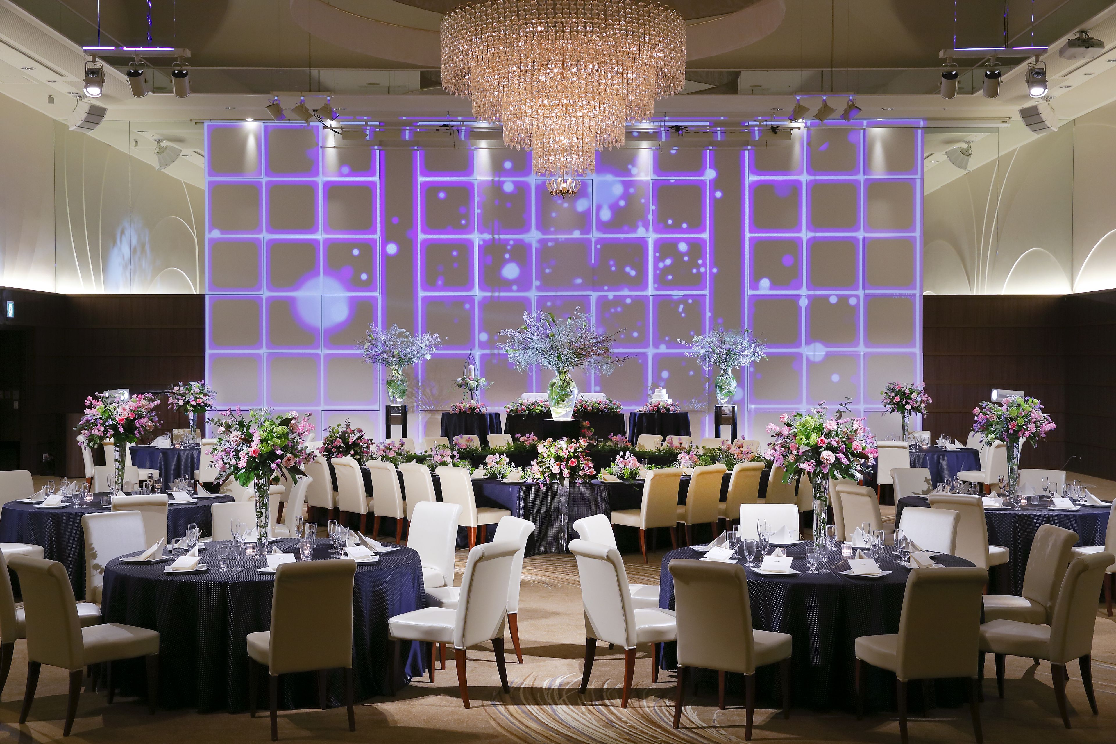 A banquet hall with round tables, white chairs, and floral centerpieces.