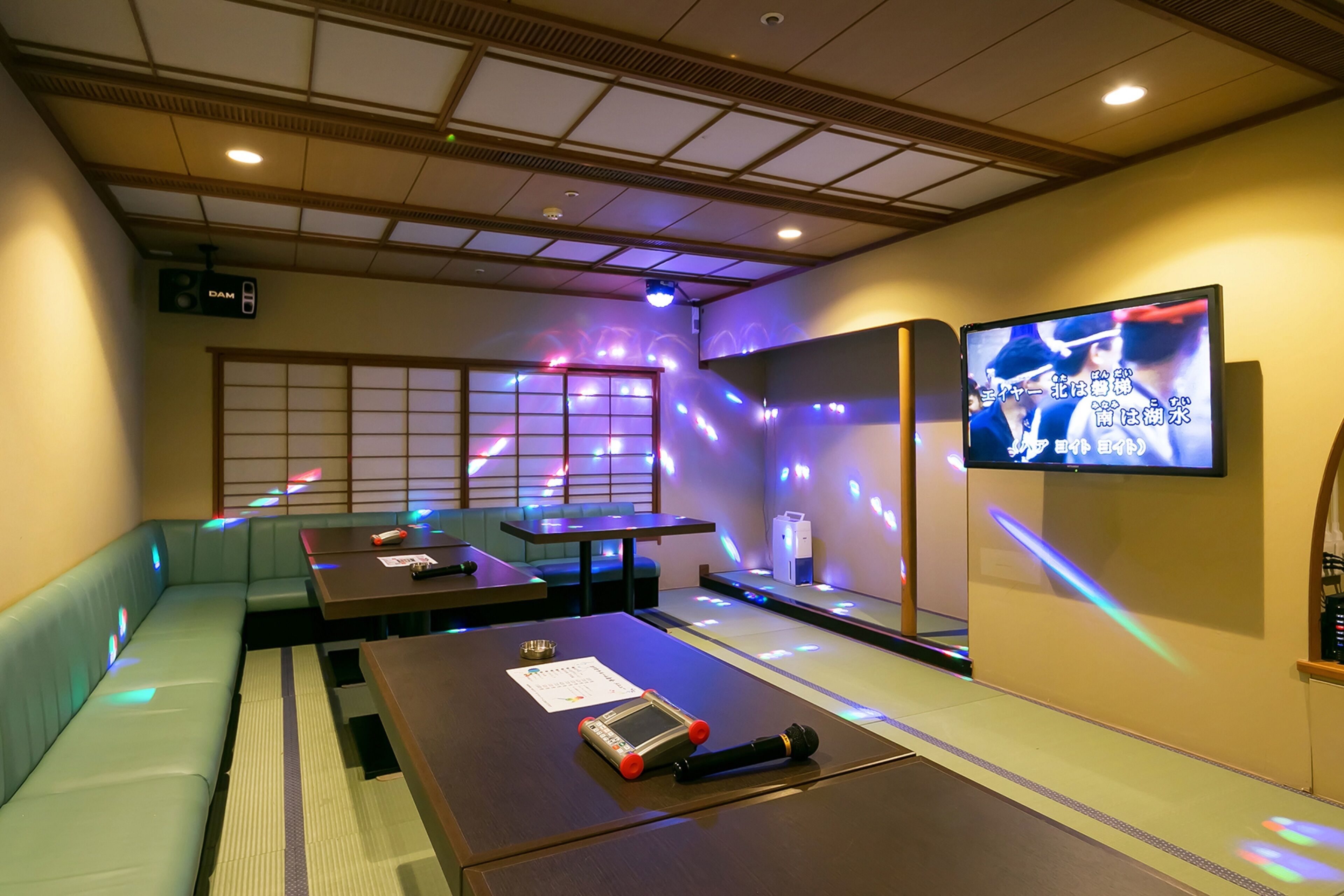 A room with a traditional Japanese aesthetic, featuring tatami flooring, low tables, and cushions. A television screen is mounted on the wall, and there is a sign indicating the room number.