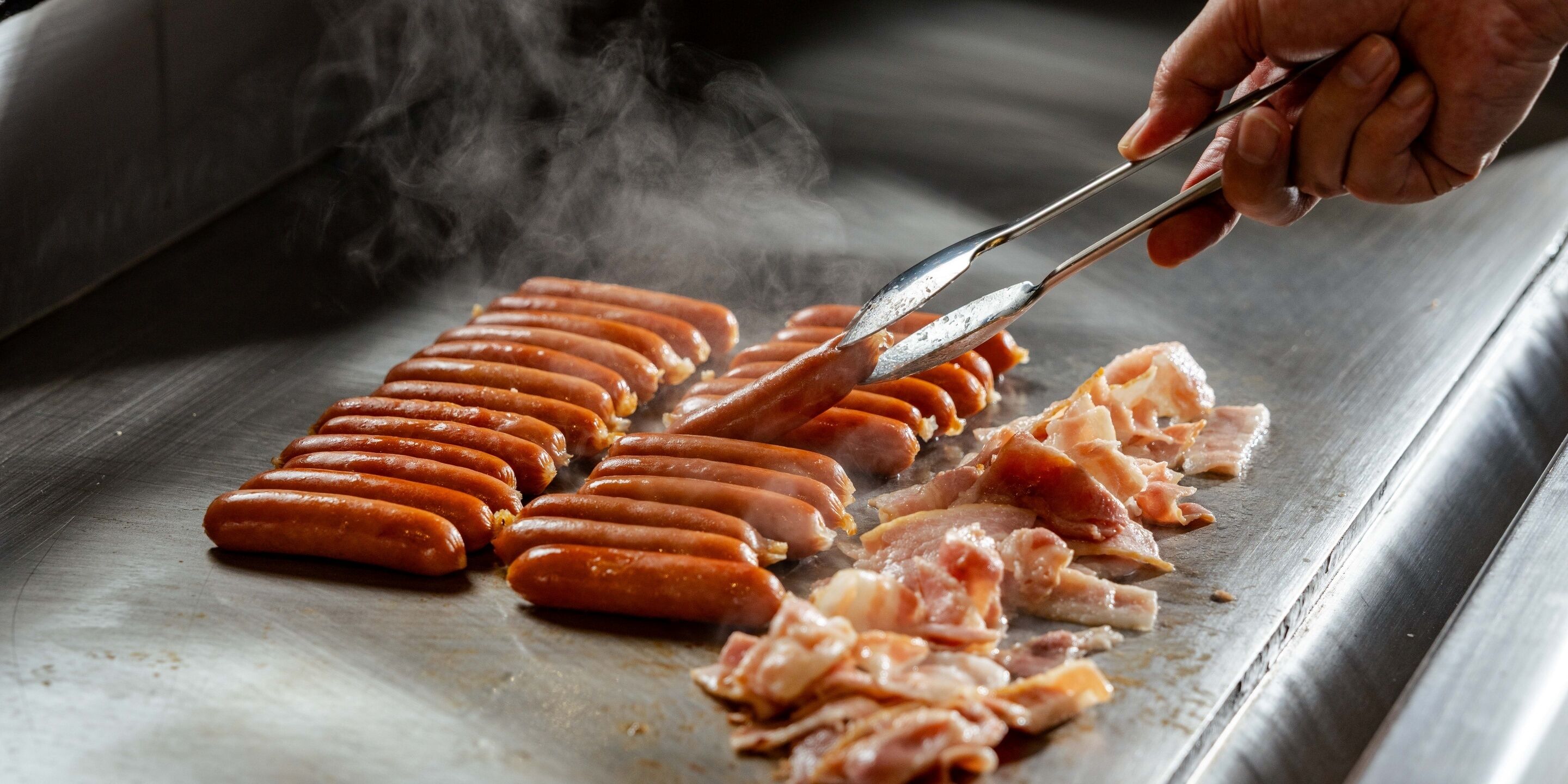 Eine Person grillt Würstchen und Fleisch auf einer Grillplatte.