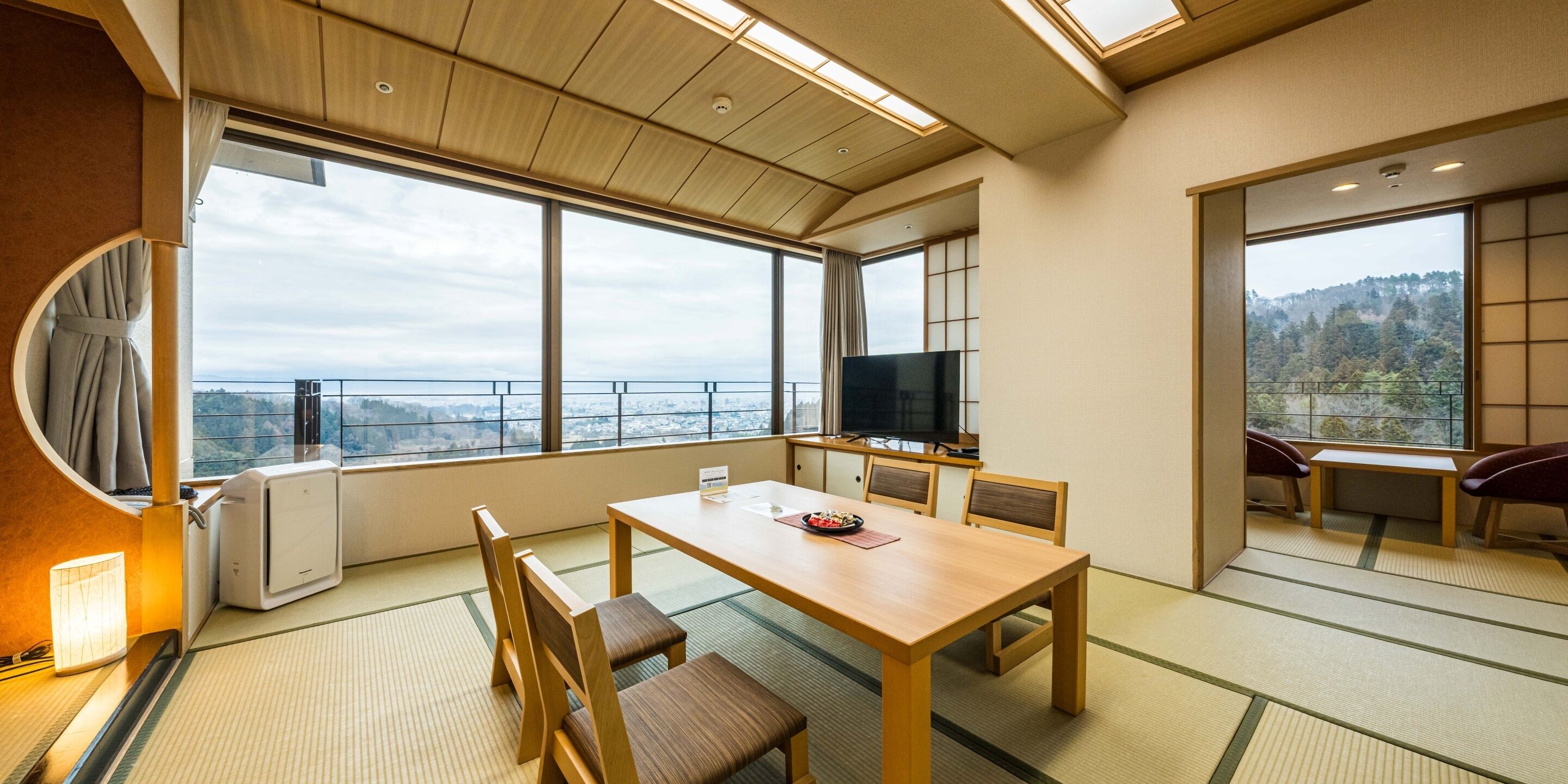 A room with tatami flooring, a wooden table, chairs, a television, and large windows offering a view of a cityscape.