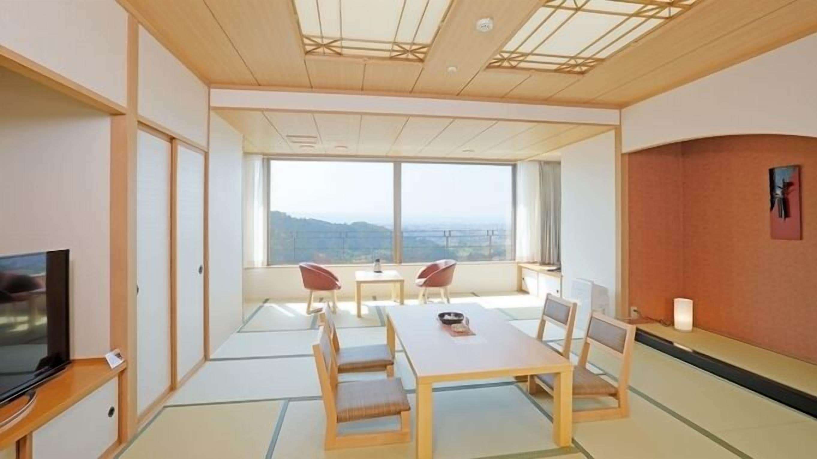 A room with tatami flooring, a television, a dining table with chairs, and a large window offering a view of the landscape.