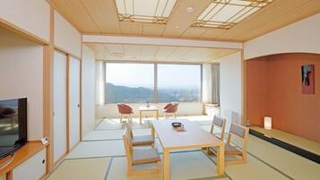 A room with tatami flooring, a television, a dining table with chairs, and a large window offering a view of the landscape.