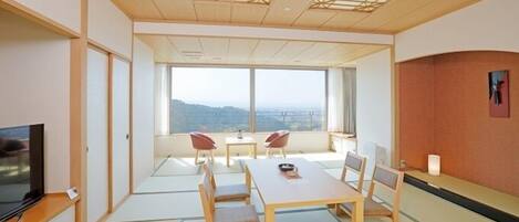 A room with tatami flooring, a television, a dining table with chairs, and a large window offering a view of the landscape.