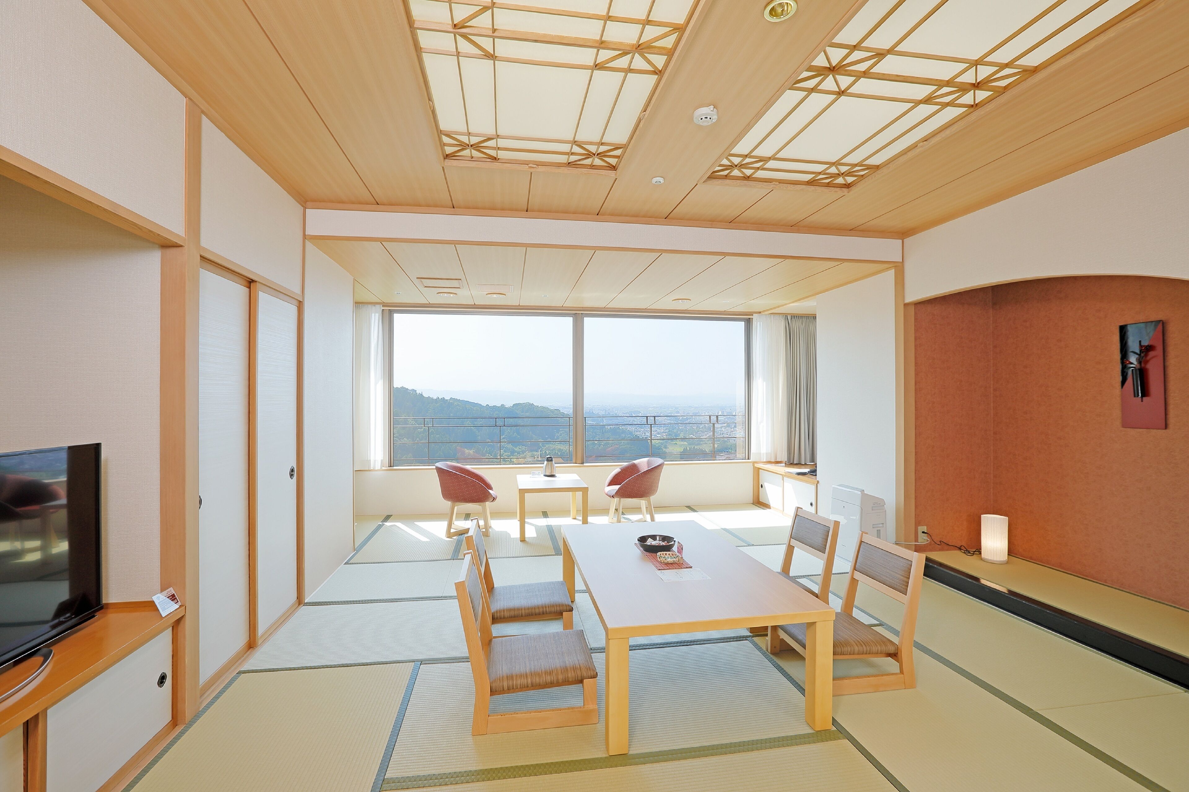 Une chambre d’hôtel moderne avec un sol recouvert de tatamis, une table à manger en bois avec des chaises, un téléviseur et une grande fenêtre donnant sur la ville.