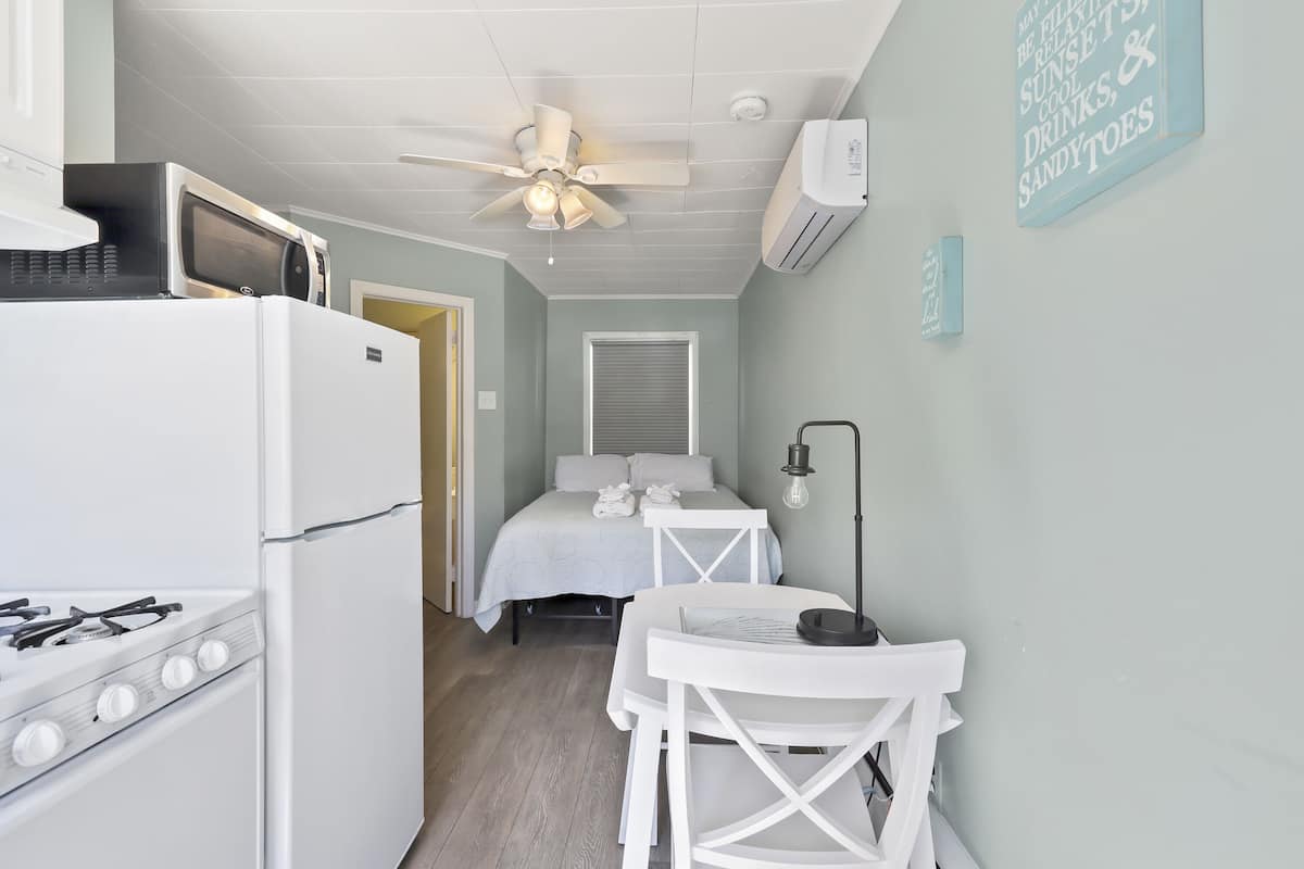 A compact kitchen with a stove, microwave, and ceiling fan. A bed with a white headboard and pillows is visible in the background.