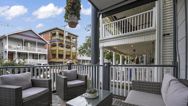 A balcony with wicker furniture and hanging plants, overlooking a street with colorful buildings. - The Inn at Ocean Grove (Ocean Grove)