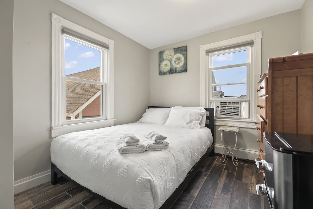 A bedroom with a bed, two windows, a painting, a heater, and a wooden cabinet.