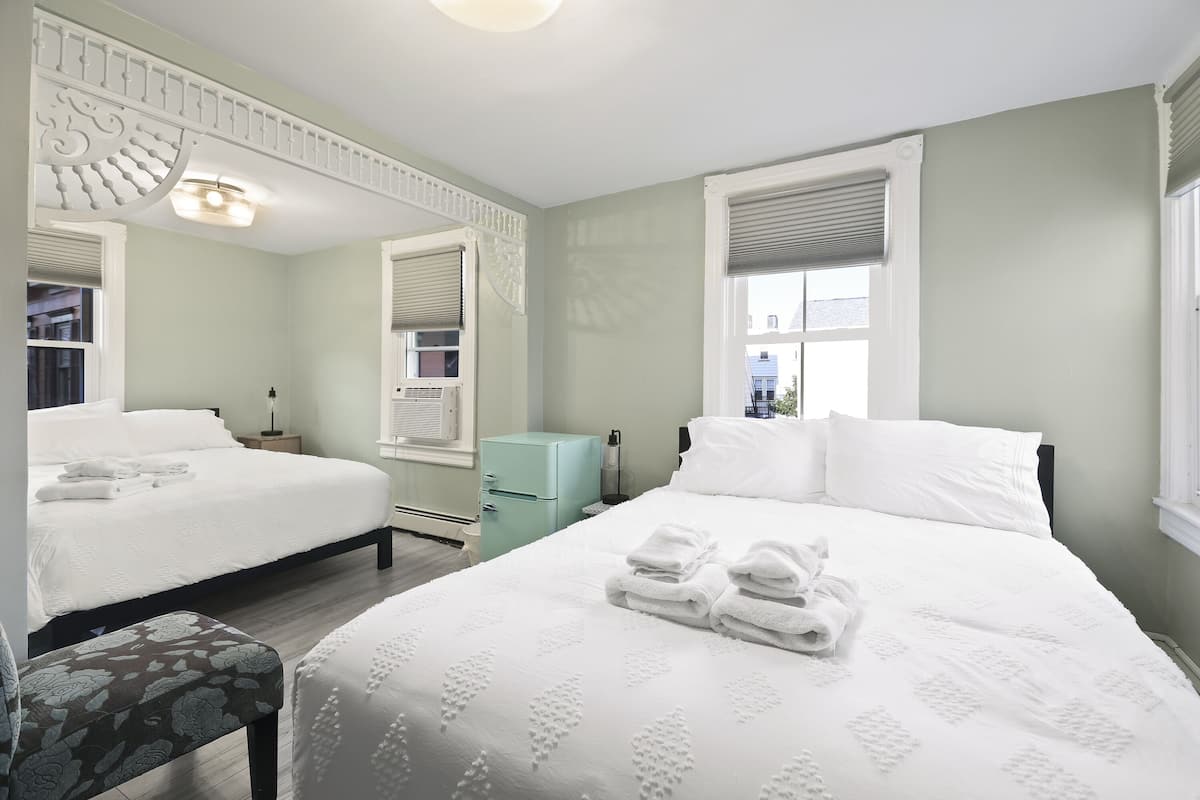 A hotel room with two beds, a small desk, a chair, and a window with blinds.