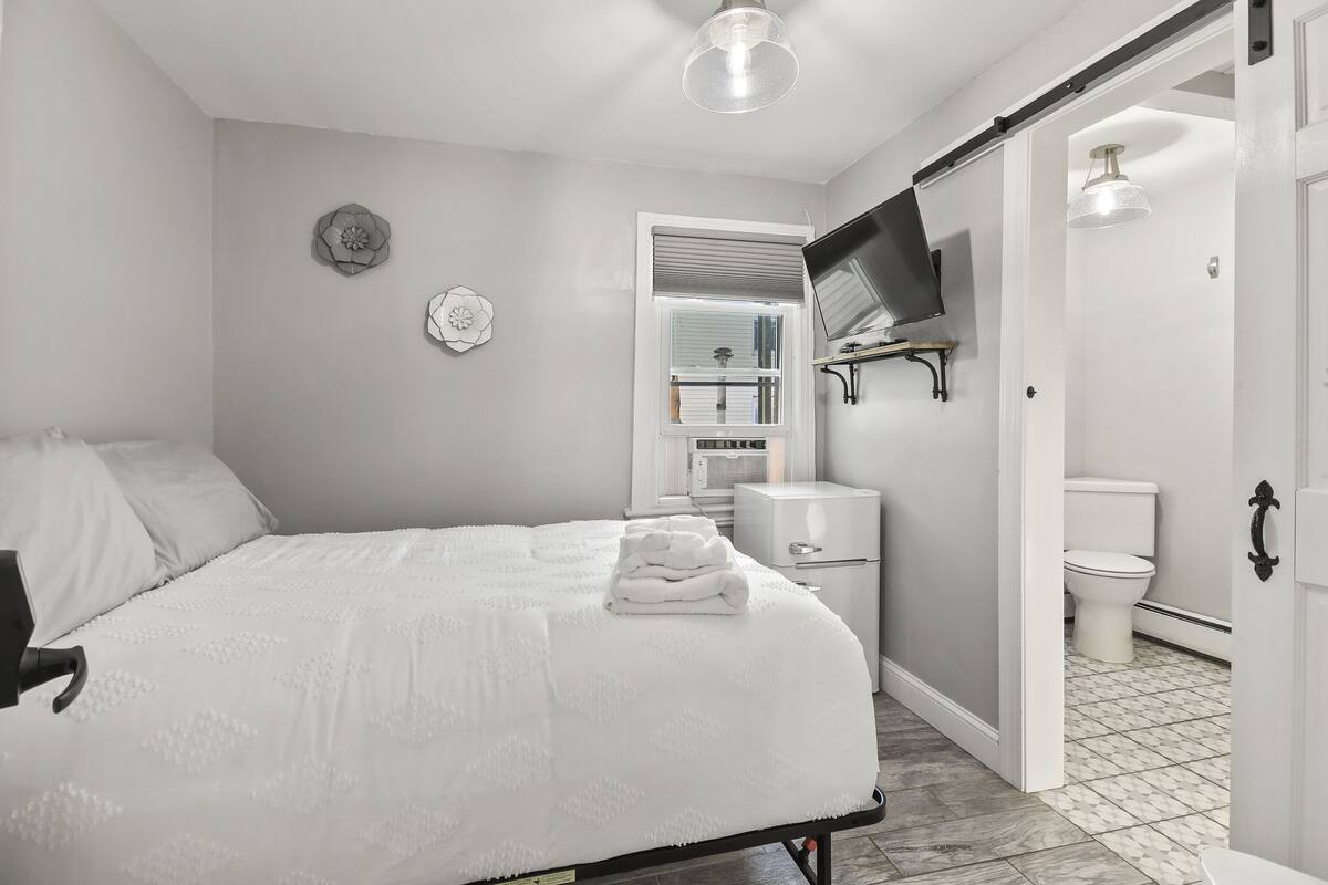 A bedroom with a bed, a TV mounted on the wall, a toilet, and a bathroom with a tiled floor.