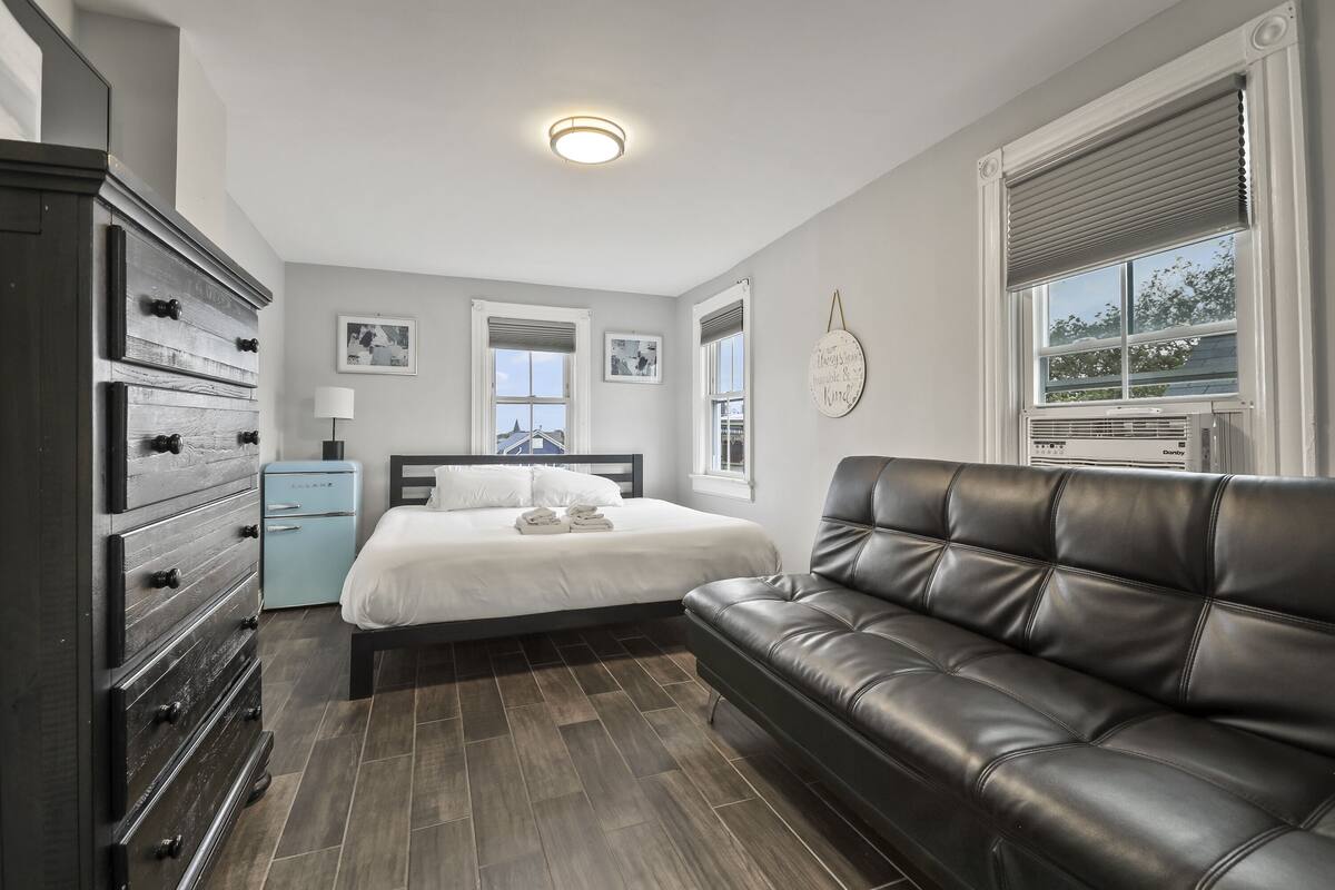 A bedroom with a bed, a dresser, a small blue appliance, a leather couch, and a window with blinds.